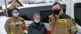 Na zdjęciu widzimy druhów ochotniczej straży pożarnej stojących po lewej i prawej stronie osoby starszej. W tle widzimy samochód oznakowany straży typu bus. 
