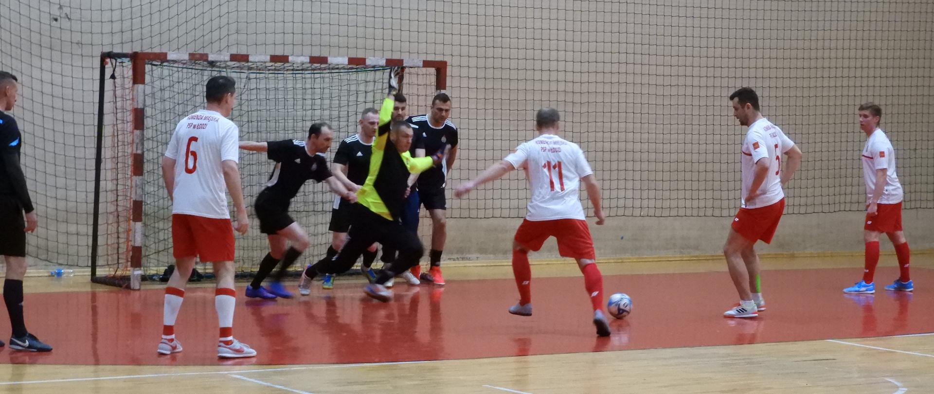 Mecz piłki nożnej w hali, zawodnicy ubrani na czarno bronią drużyny ubranej na biało i czerwono, próbując zdobyć bramkę blisko bramki.