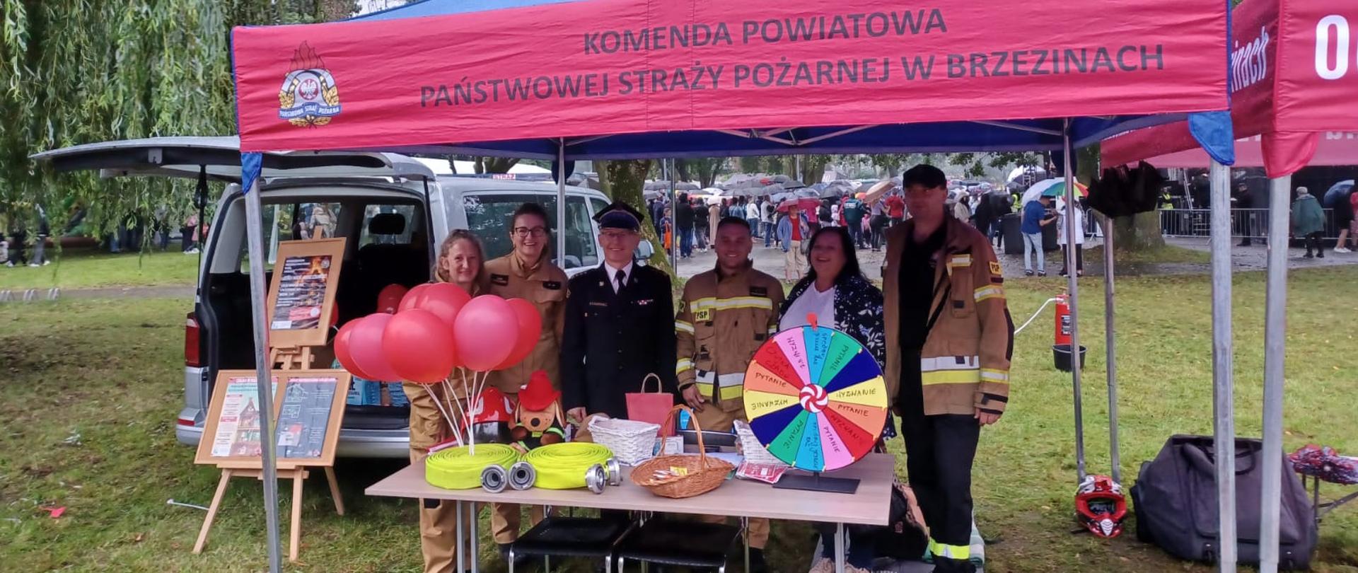 Funkcjonariusze i pracownicy cywilni stoją pod czerwonym namiotem podczas festiwalu i prowadzonej akcji Czad i Ogień. Obudź czujność. Na stołach przed nimi porozkładane są czujki tlenku węgla oraz materiały promocyjne. Za namiotem widoczny samochód typu BUS straży pożarnej, w oddali za namiotem widać tłum ludzi z parasolami.