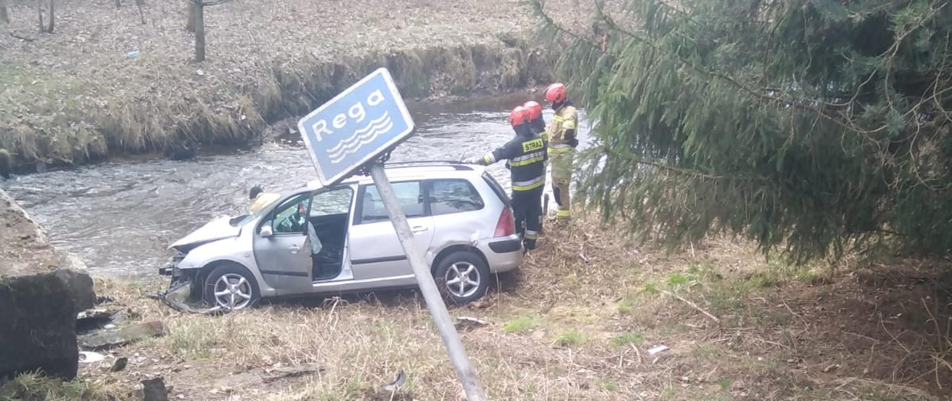 Zdjęcie przedstawia rzekę oraz uszkodzony znak z napisem rzeki Rega. Przed rzeką stoi rozbite srebrne auto. Brakuję 1 m aby auto znalazło się w rzece. Obok auta znajdują się strażacy.