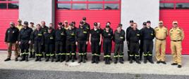 Strażacy oraz członkowie komisji egzaminacyjnej stojący przed budynkiem w dwuszeregu - pozujący do zdjęcia grupowego.