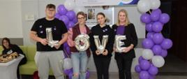 Kilkoro uczniów z nauczycielką pozują do zdjęcia trzymając balony układające się w napis LOVE, obok fioletowe balony