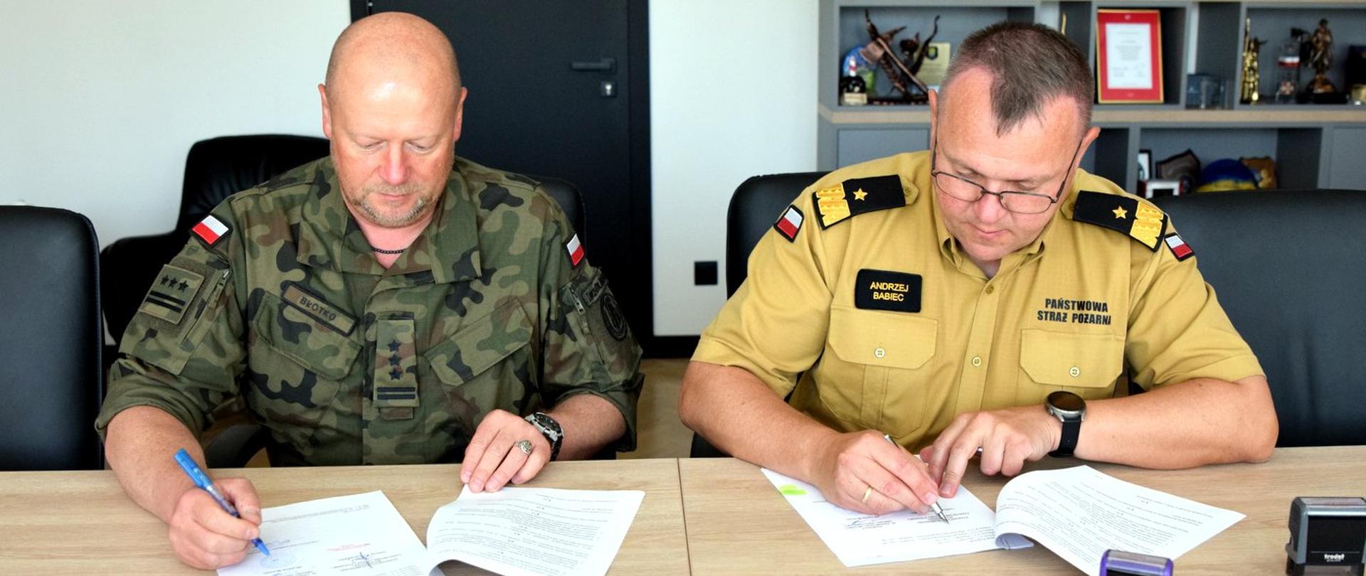 Zdjęcie zrobione w pomieszczeniu. Przy stole siedzą od lewej strony: pułkownik Wojsk Obrony Terytorialnej i oficer Państwowej Straży Pożarnej w stopniu generała. Oficerowie podpisują dokumenty. 