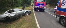 Zdjęcie przedstawia rozbity samochód osobowy marki VW Passat znajdujący się w przydrożnym rowie oraz samochód straży pożarnej, pogotowia i policji znajdujące się na jezdni. 