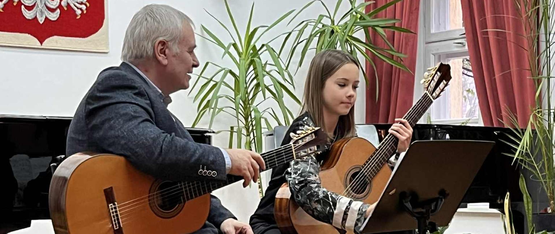 Dwójka osób siedzi na scenie i gra na gitarach. Po lewej stronie siedzi dr hab. Jacek Dulikowski, a po prawej uczennica. Oboje trzymają gitary i patrzą na nuty ustawione na pulpicie. W tle widać zielone rośliny, oraz godło Polski.