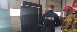 Policjant w ciemnoniebieskim mundurze i pasie taktycznym ćwiczy wyważanie drzwi za pomocą narzędzia typu Halligan. Stoi tyłem do obiektywu, skierowany w stronę specjalnej makiety treningowej w garażu remizy strażackiej. Po prawej stronie przygląda się strażak w pełnym umundurowaniu bojowym.