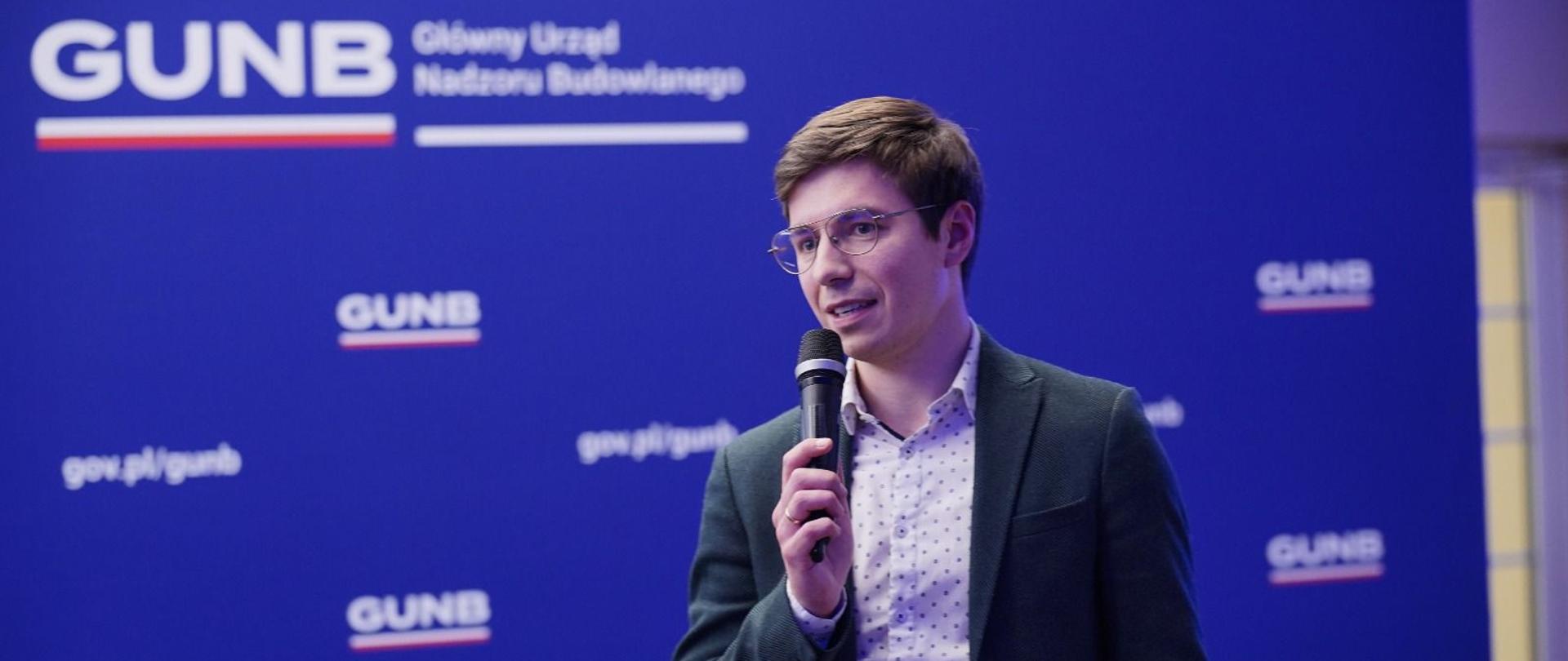 Filip Prusik-Serbinowski, Zastępca GINB podczas wystąpienia na spotkaniu roboczym FEnIKS, w tle granatowa ścianka z logotypami GUNB 