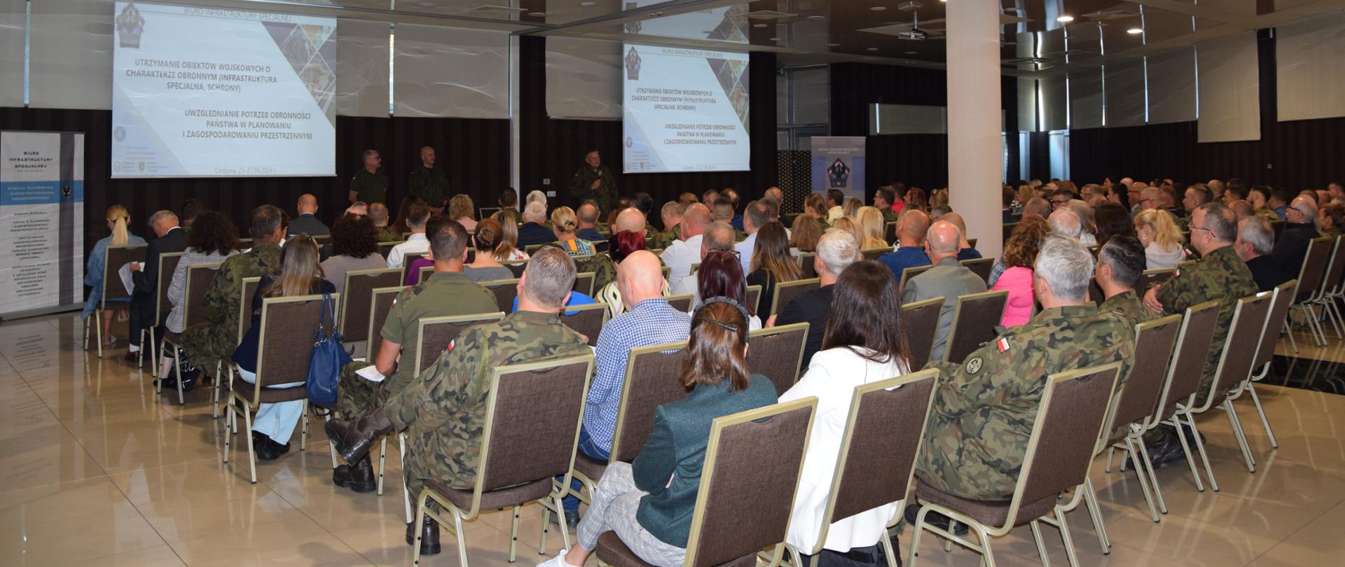 Sala kinowa w biało-beżowych odcieniach. Na pierwszym planie widać ludzi siedzących na krzesłach. W oddali stoi mężczyzna na scenie. Na środku sali wiszą ekrany projekcyjne, na których wyświetlana jest prezentacja.