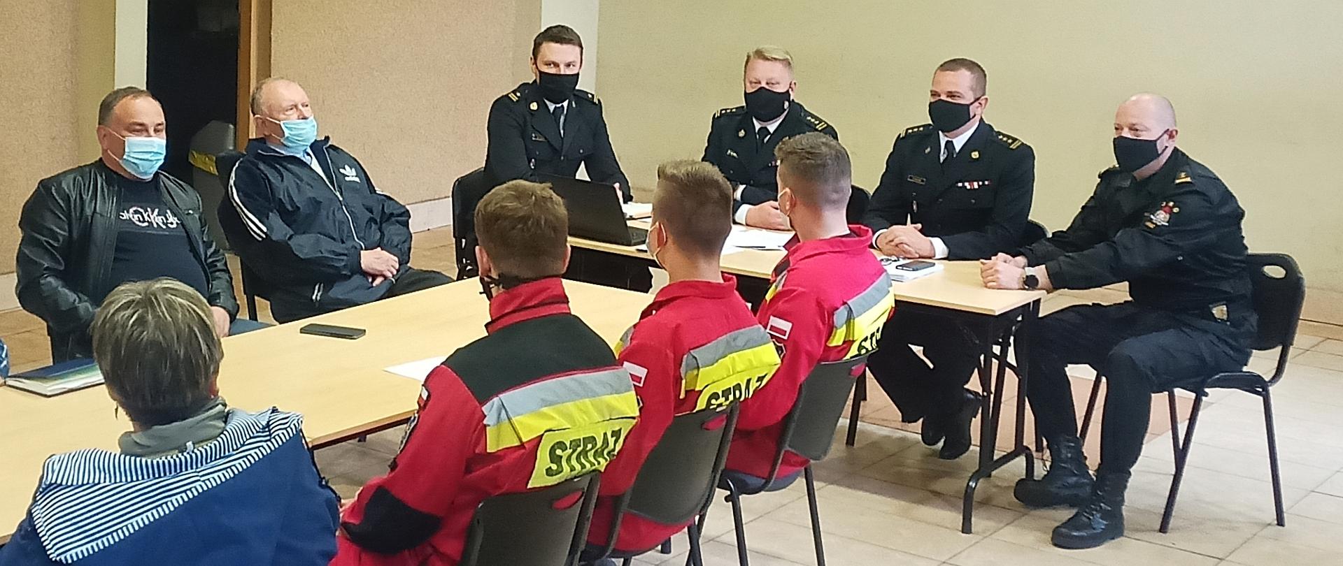Na zdjęciu widać 4 strażaków PSP w mundurach galowych, 3 strażaków w czerwonych kombinezonach oraz 3 osoby w normalnych ubraniach siedzących przy dwóch stołach w czasie spotkania