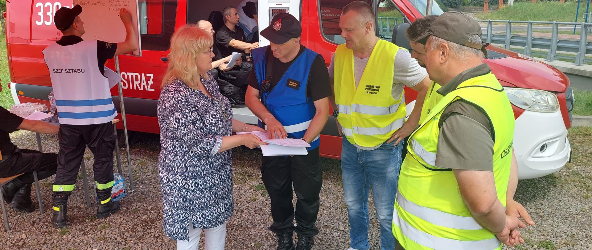 Praca sztabu w namiocie, od lewej strony szef sztabu w białej kamizelce notuje czarnym flamastrem na białej tablicy, z prawej strony zespół 4 osób spogląda na dokumentacje, którą trzyma funkcjonariusz PSP w niebieskiej kamizelce, pozostali w żółtych kamizelkach. W tle czerwony oznakowany pojazd pożarniczy dowodzenia i łączności w którym pracuje dwóch funkcjonariuszy PSP.