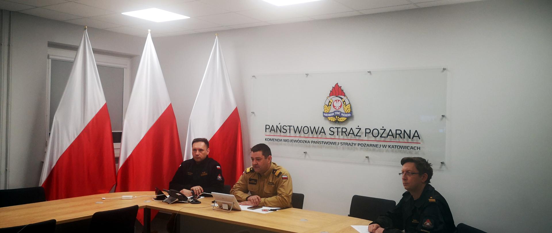 Na zdjęciu sala sztabowa. Śląski Komendant Wojewódzki PSP prowadzi wideokonferencję z kadrą kierowniczą PSP woj. śląskiego. Po lewej stronie siedzi Naczelnik Wydziału Planowania i Działań Ratowniczych. W tle po lewej stronie ustawione trzy flagi narodowe. Na ścianie napis Państwowa Straż Pożarna i logo PSP.