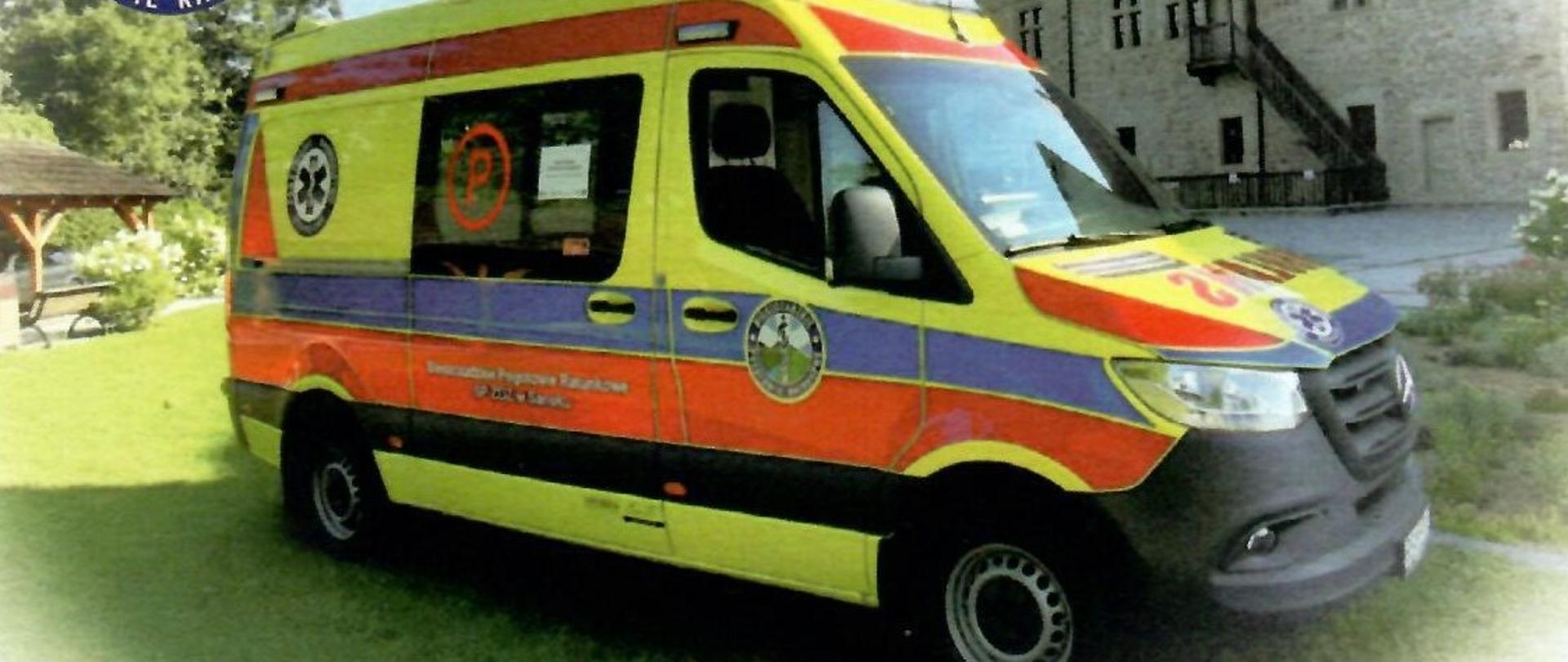 Na zdjęciu widać karetkę Pogotowia Ratunkowego w żółto-niebiesko-czerwonych barwach stojącą pod budynkiem na trawie.
