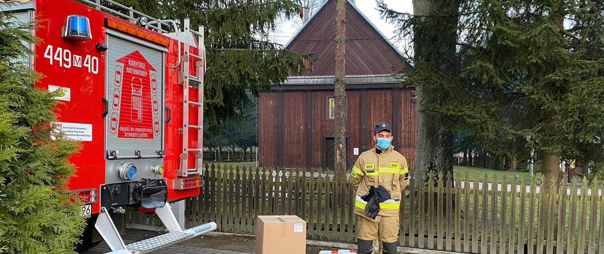 Obok samochodu pożarniczego i ułożonych kartonów z maseczkami i pojemników z płynem dezynfekcyjnym stoi strażak w ubraniu specjalnym. W tle widać za drzewami widać drewniany kościół.