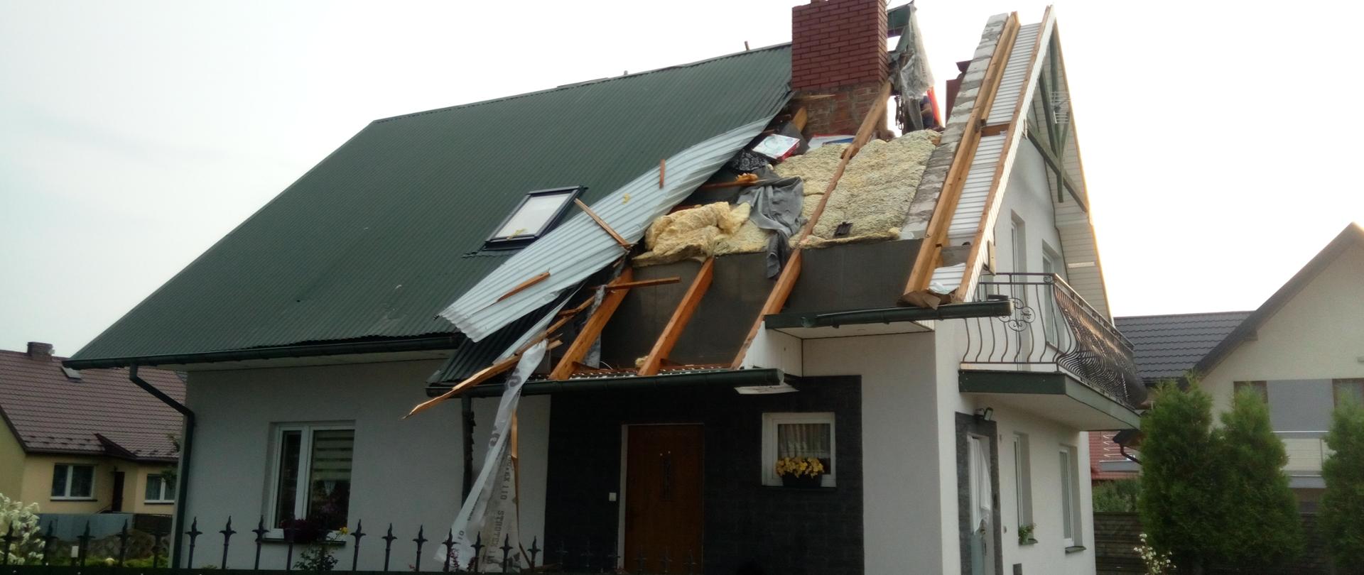 Dom jednorodzinny uszkodzony w wyniku silnego wiatru. Zerwana została znaczna część pokrycia dachowego z blachy odsłaniająca konstrukcję dachową wraz z warstwami izolacyjnymi.