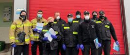Dystrybucja ulotek. Dziewięciu strażaków stojących na tle strażnicy OSP.. Strażacy trzymają w ręku ulotki.