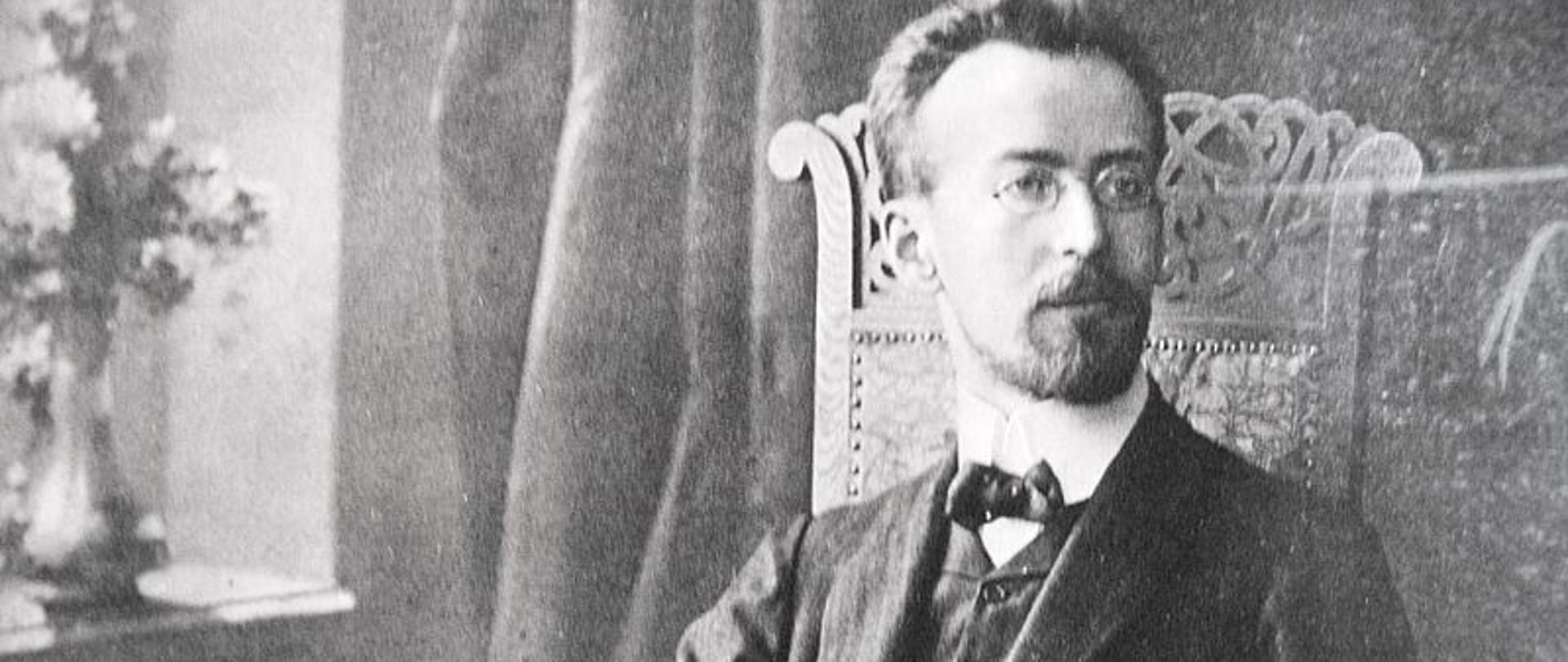 zdjęcie przedstawia patrol szkoły, Mieczysława Karłowicza siedzącego na drewnianym fotelu ze zdobieniem na oparciu, jego lewa ręka jest oparta na podłokietniku, w prawej, wspartej na stoliczku trzyma notatnik, głowa muzyka zwrócona jest w lewą stronę, w tle znajduje się okno z flakonem kwiatów na parapecie