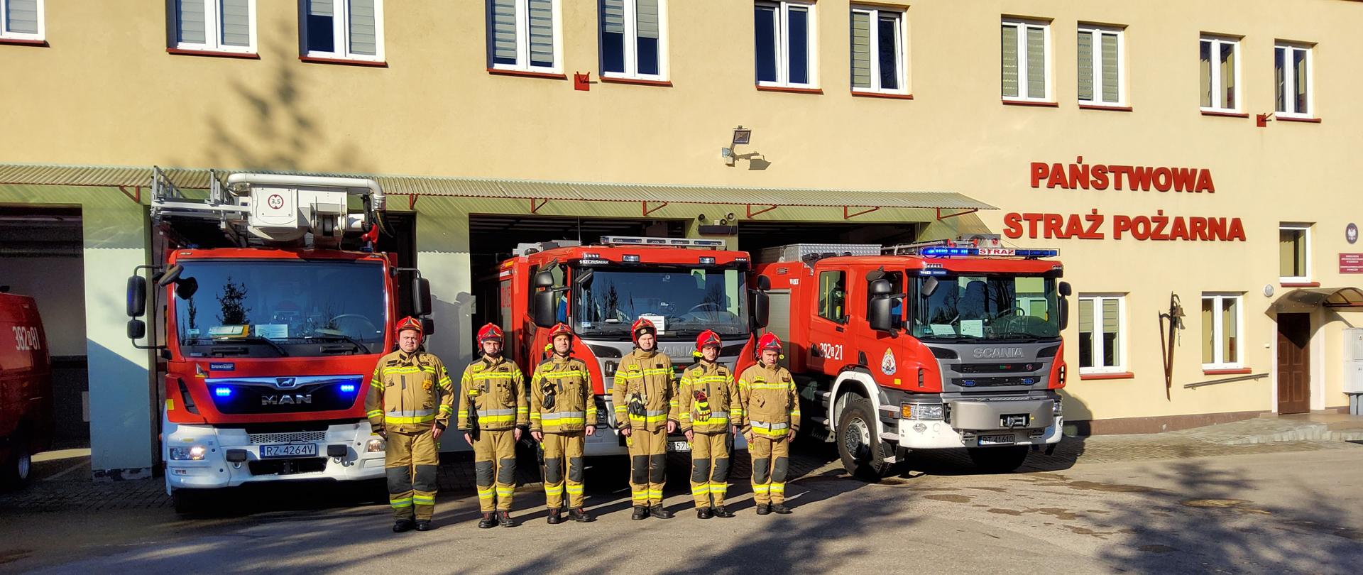 Strażacy stoją na zbiórce u umundurowaniu bojowym na placu przed budynkiem remizy. Za nimi widoczne ustawione pojazdy gaśnicze.