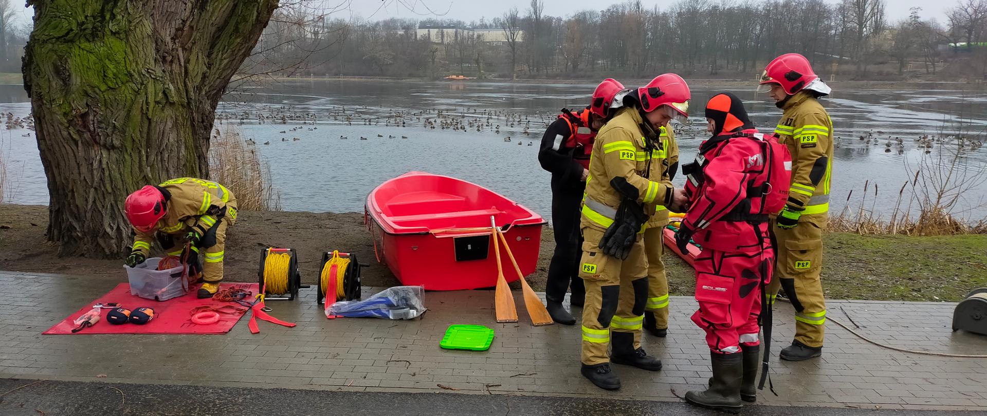 Na zdjęciu widać strażaków stojących nad brzegiem jeziora, którzy przygotowują sprzęt do ćwiczeń z ratownictwa lodowego