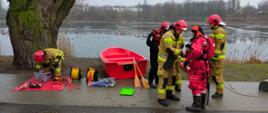 Na zdjęciu widać strażaków stojących nad brzegiem jeziora, którzy przygotowują sprzęt do ćwiczeń z ratownictwa lodowego
