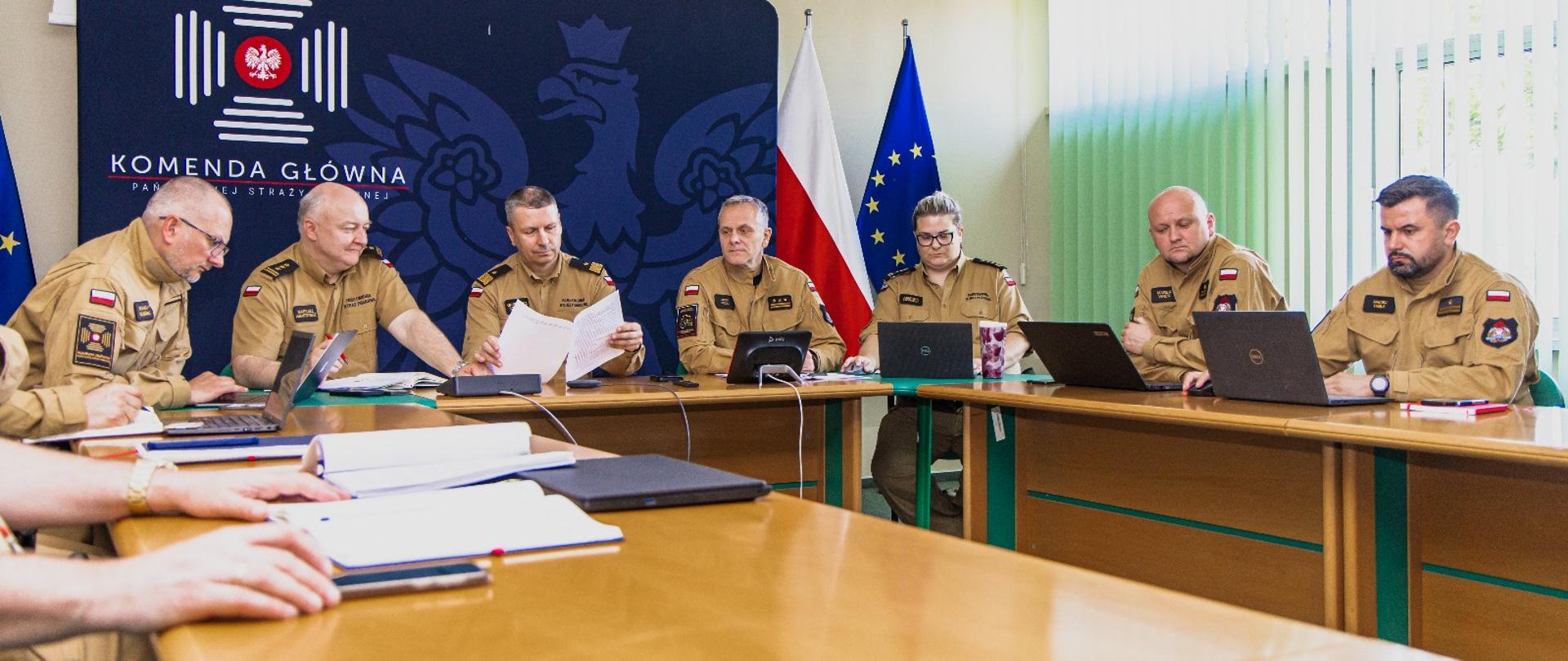 Uczestnicy odprawy w KG PSP przeglądają dokumenty oraz omawiają treści wdrażanych rozwiązań prawnych. Po prawej stronie kadru widać skupienie na pracy i przygotowaniach do wystąpień.
