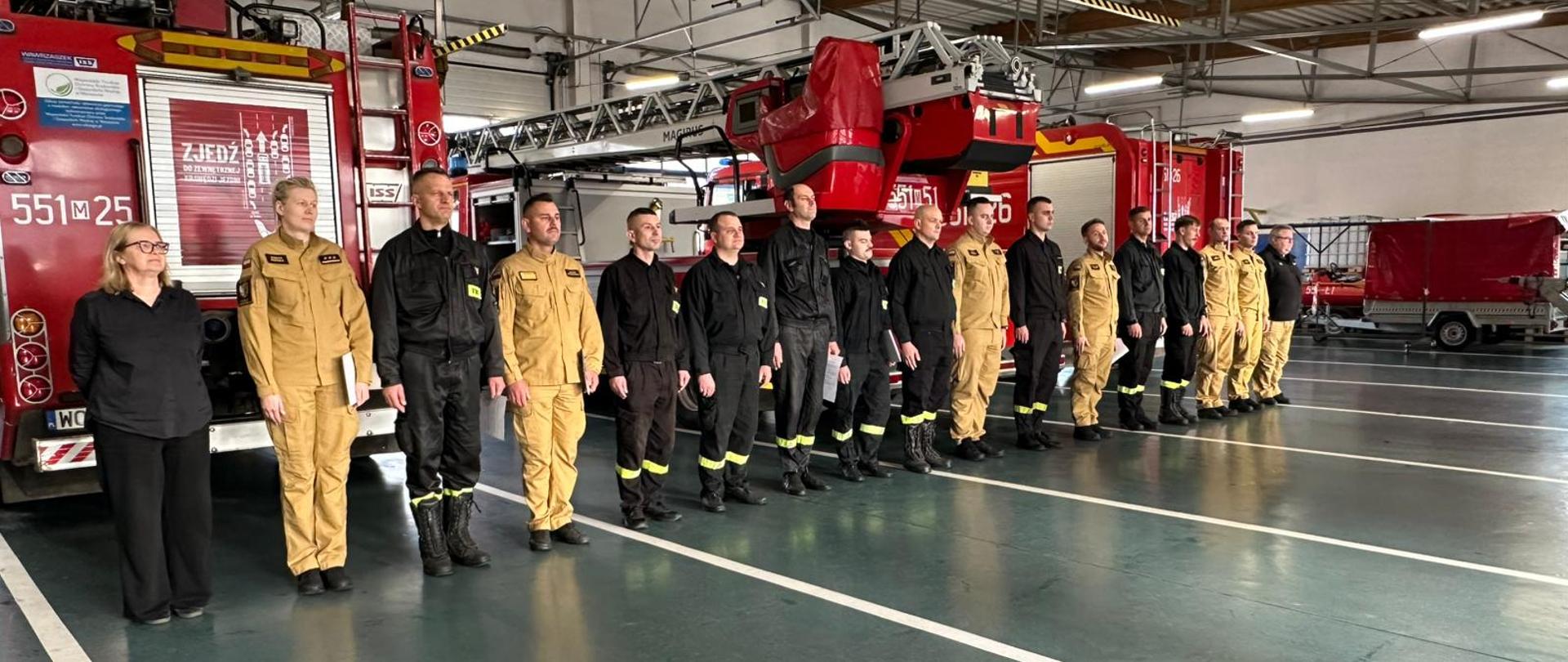 Zdjęcie przedstawia uroczystą scenę wewnątrz remizy strażackiej, gdzie grupa strażaków stoi w równym szeregu przed czerwonymi wozami strażackimi. W tle widać wysokie sufity z odsłoniętymi belkami, jasne oświetlenie oraz wypolerowaną podłogę, co podkreśla porządek i profesjonalizm miejsca.
👨‍🚒 Strażacy:
• Ubrani są w dwa typy mundurów: czarne z żółtymi pasami odblaskowymi oraz beżowe, typowe dla odzieży ochronnej używanej podczas akcji.
• Ich postawa jest formalna i zdyscyplinowana, co sugeruje, że uczestniczą w apelu, inspekcji lub ceremonii.
🚒 Wóz strażacki:
• W tle widoczne są pojazdy ratownicze wyposażone w drabiny i sprzęt gaśniczy.
• Ich obecność podkreśla gotowość operacyjną jednostki.
👩 Kobieta po lewej stronie:
• Ubrana w czarne ubranie, stoi nieco z boku, co może wskazywać na rolę organizacyjną, gościa honorowego lub przedstawicielkę administracji.
