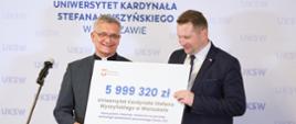 Minister Czarnek stoi obok starszego mężczyzny w garniturze, trzymają duży symboliczny czek z napisem 5 999 320 zł, za nimi na ścianie napis UKSW.