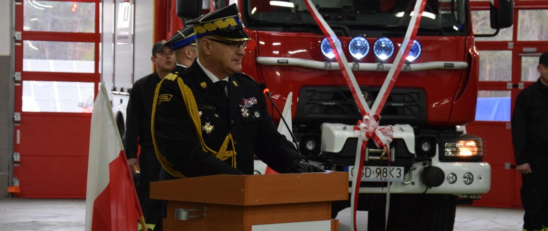 Przy mównicy ustawionej w garażu stoi komendant wojewódzki w mundurze galowym. Za nim udekorowany pojazd pożarniczy.