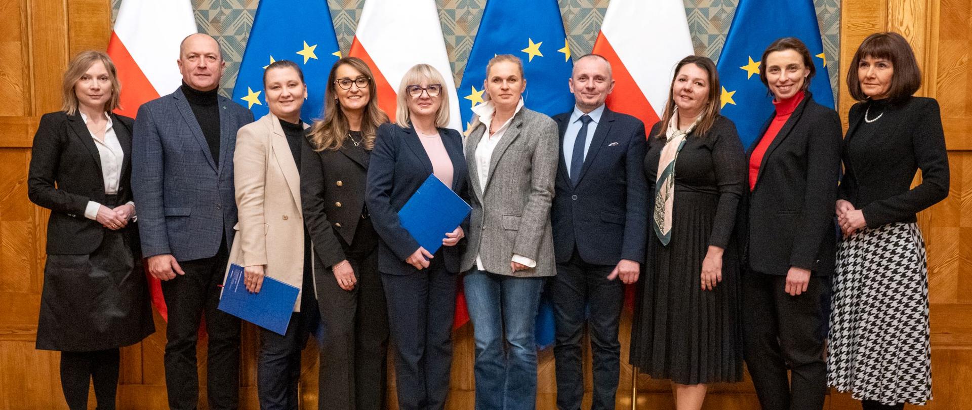 Grupa kobiet i mężczyzn pozuje do zdjęcia. W tle trzy flagi biało-czerwone i trzy flagi niebieskie z żółtymi gwiazdkami.