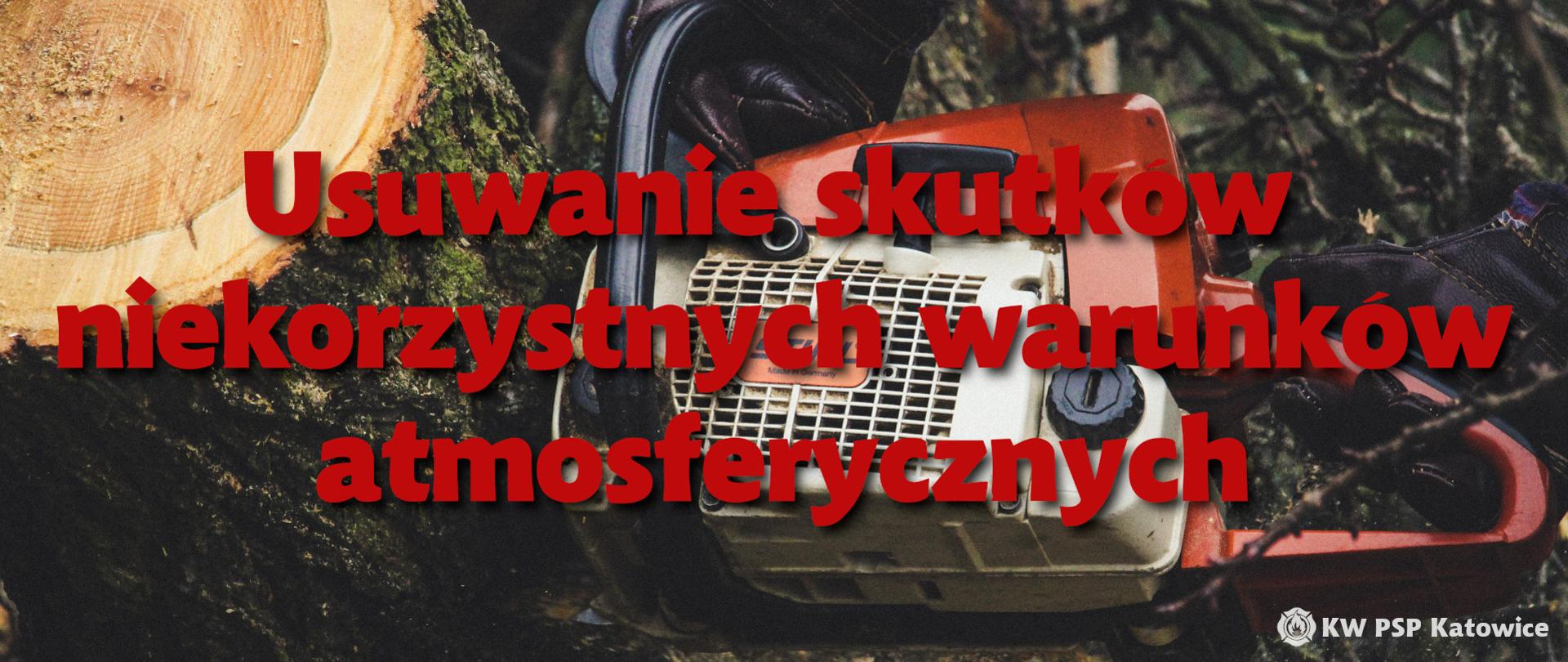 Zdjęcie przedstawia czerwony napis usuwanie skutków niekorzystnych warunków atmosferycznych, w tle fragment spalinowej pilarki do drzewa i ściętego pnia drzewa. W prawym dolnym rogu napis KW PSP Katowice.