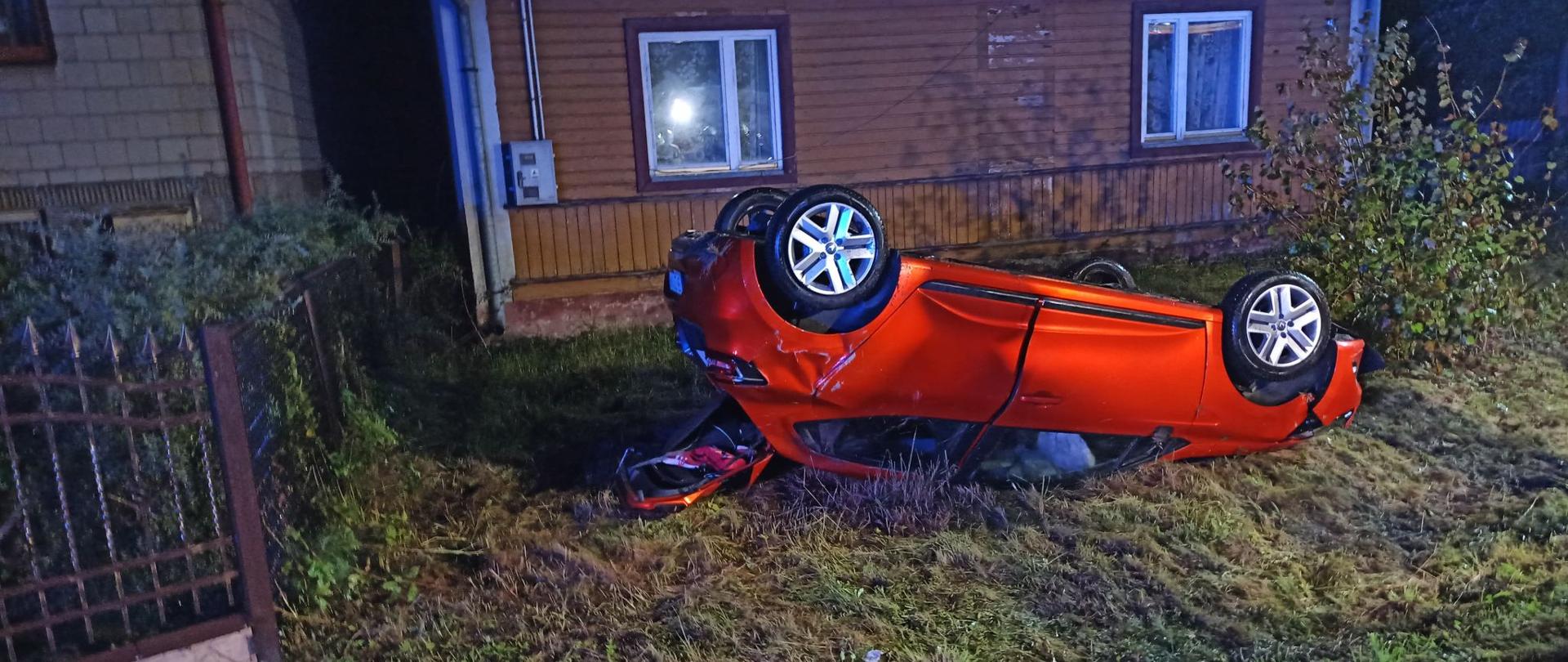 Czerwony samochód osobowy leży przewrócony na dach przed posesją domu jednorodzinnego 