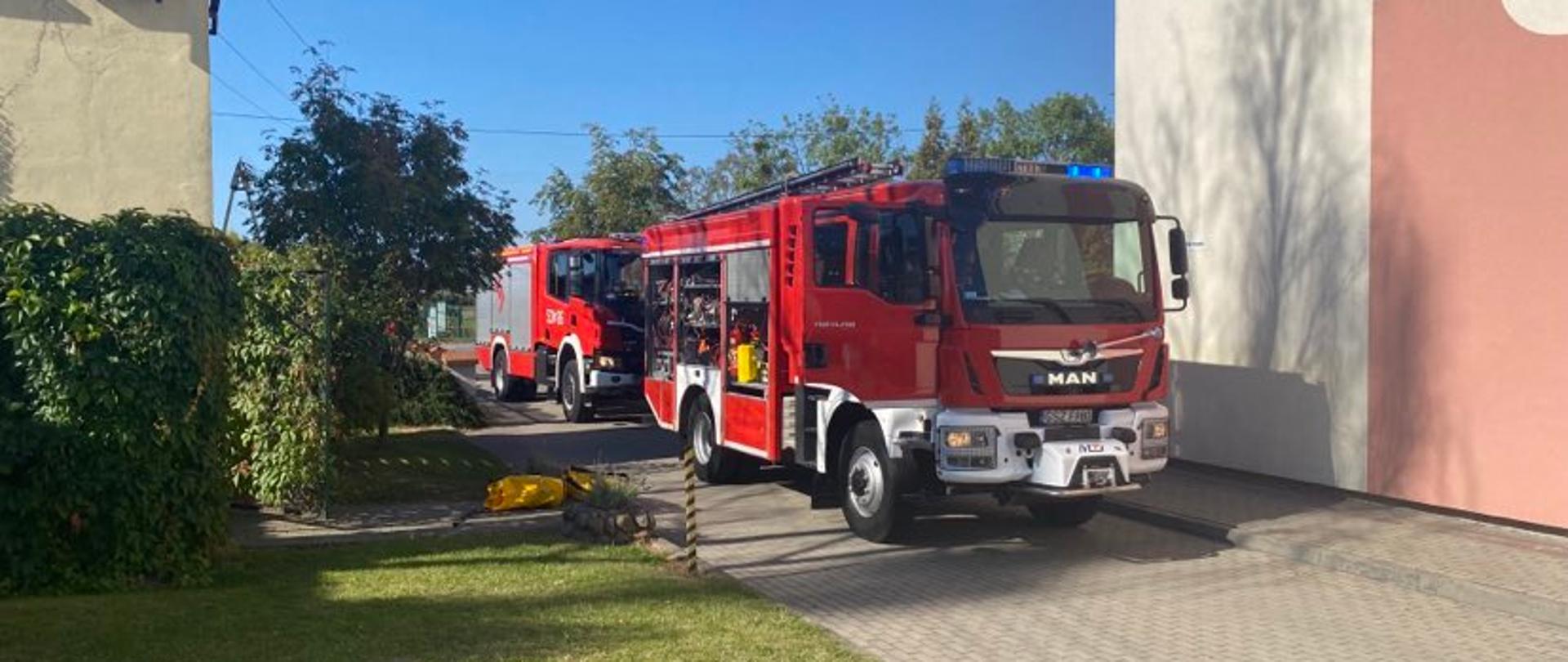 kadr przedstawia dwa czerwone pojazdy pożarnicze stojące w pobliżu szkoły.