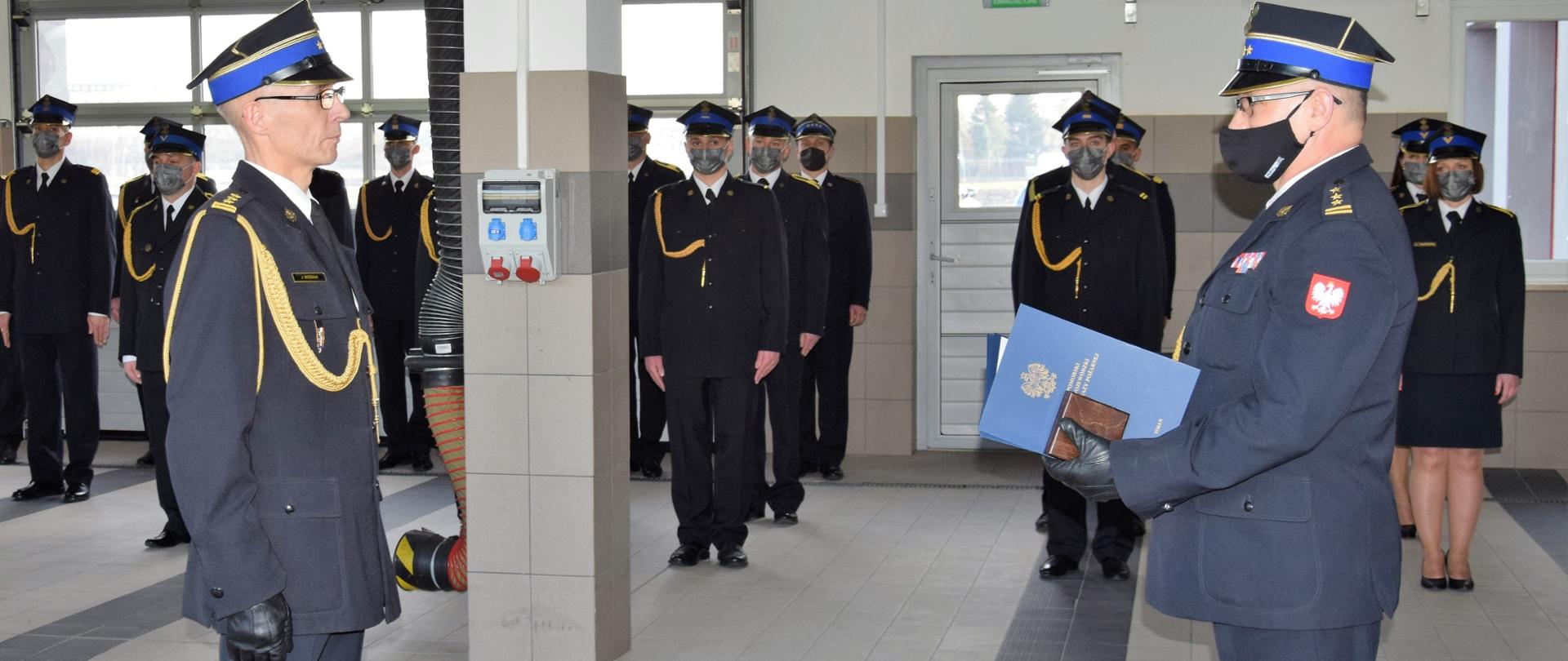 Na zdjęciu stoi komendant powiatowy PSP w Wąbrzeźnie, który odbiera od komendanta wojewódzkiego PSP decyzję zwalniającą ze służby. W tle pracownicy KP PSP w Wąbrzeźnie. 