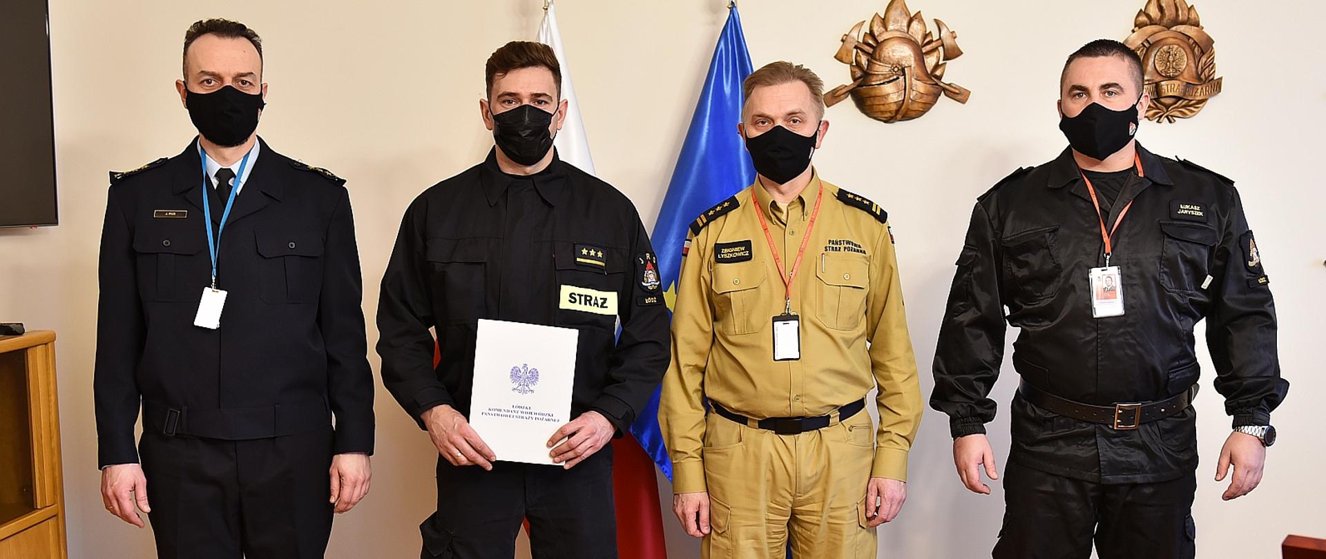 na zdjęciu czterech strażaków w czterech rodzajach umundurowania od lewej strażak w umundurowaniu służbowym starego typu, strażak w ubraniu koszarowym, strażak w umundurowaniu służbowym nowego typu oraz strażak w umundurowaniu dowódczo sztabowym, strażak w umundurowaniu koszarowym trzyma w rękach teczkę z decyzją, za strażakami flagi polski i uni europejskiej