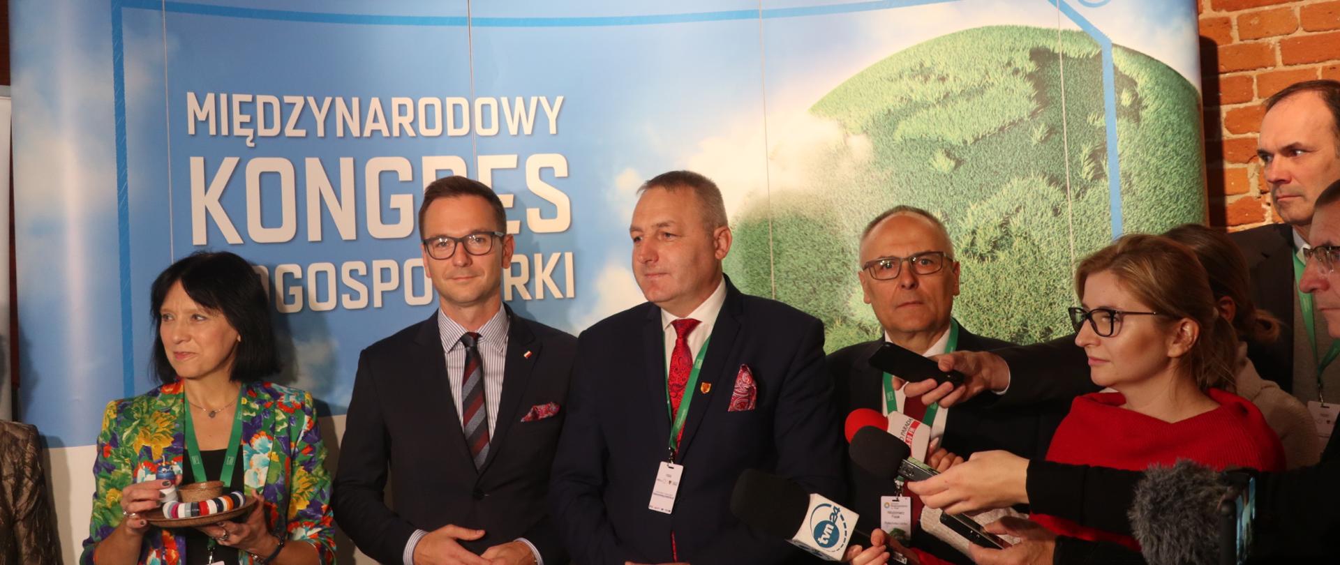 Na zdjęciu stoi sześć osób, drugi z lewej wiceminister Waldemar Buda, w tle baner z napisem Międzynarodowy Kongres Biogospodarki
