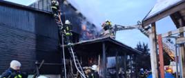Zdjęcie przedstawia pojazdy pożarnicze oraz strażaków podczas działań ratowniczo-gaśniczych na tle budynku objętego pożarem