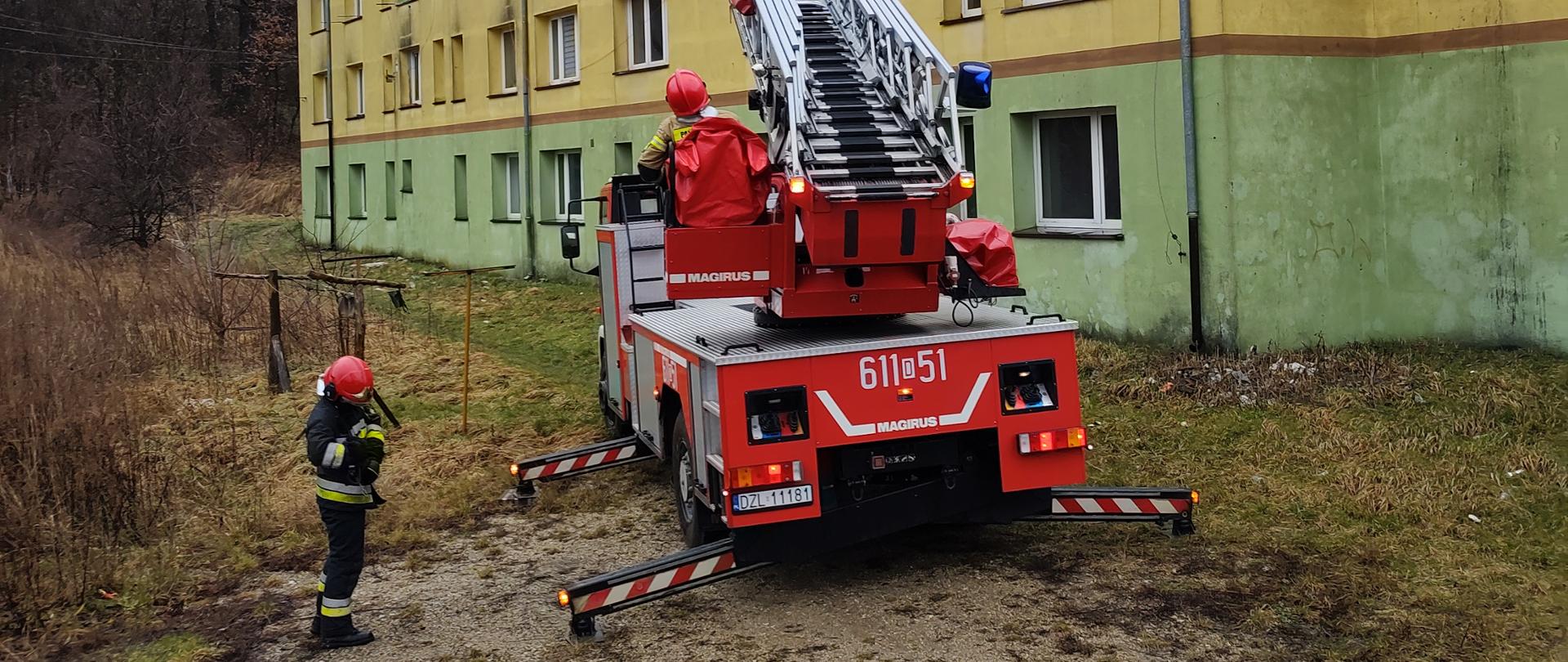 strażacki samochód typu drabina stoi na tle budynku mieszkalnego wielorodzinnego, wokół widoczni strażacy