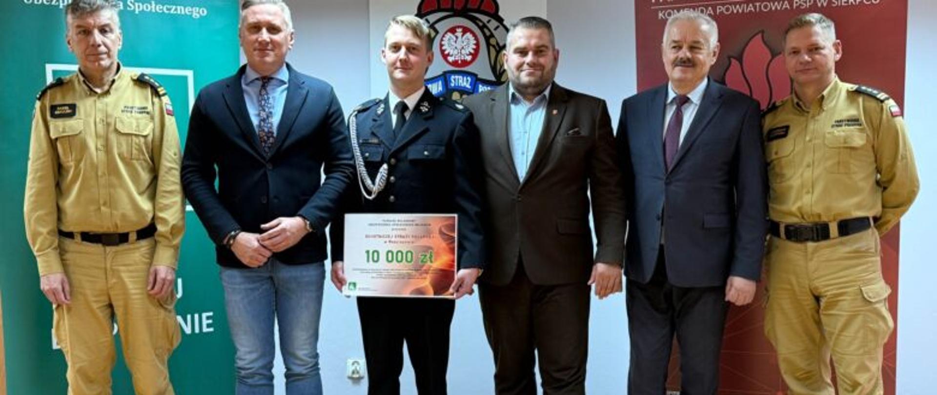 Sześciu mężczyzn stoi tle banerów KRUS i Komendy Powiatowej PSP w Sierpcu jeden z nich w galowym mundurze strażaka trzyma plansze z kwotą otrzymanego wsparcia
