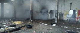 Zdjęcie wykonane wewnątrz pomieszczenia ze środka dużej opuszczonej hali. Pożarem objęta jest jego mniejsza część. Z miejsca gdzie palą się opony i śmieci wydostaje się dym. Ściany pomieszczenia pokryte graffiti. Tynk jest obity i zniszczony