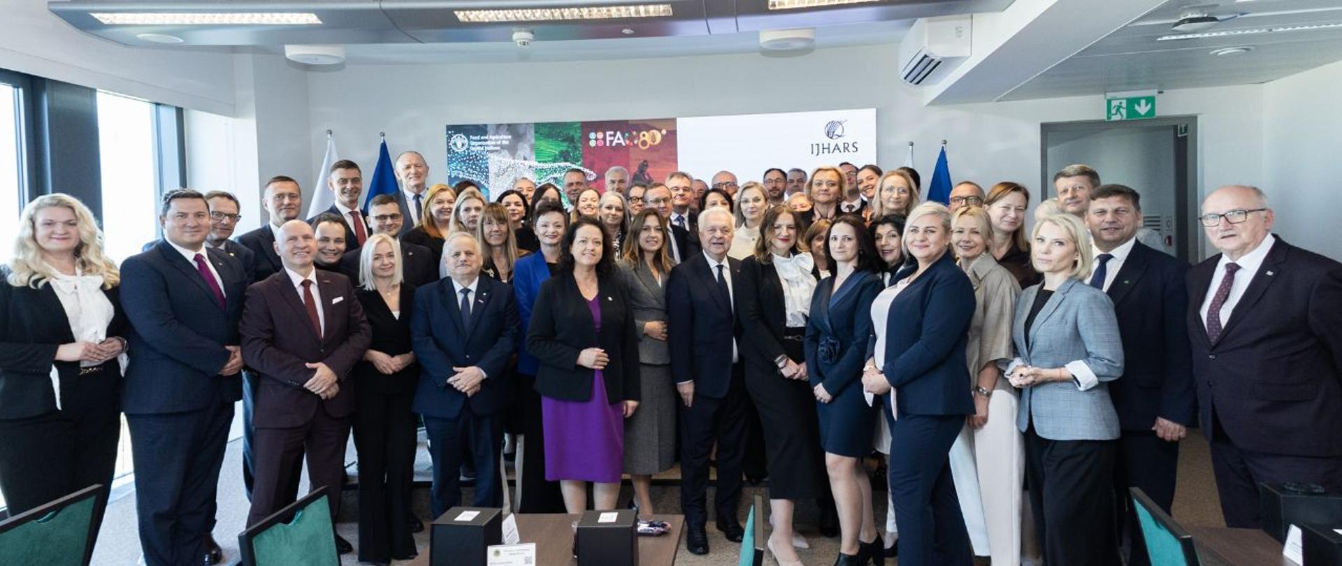 Zdjęcie grupowe wykonane w sali konferencyjnej. Kilkadziesiąt osób w strojach formalnych pozuje do wspólnej fotografii. W tle widoczne są roll-upy z logotypami FAO i IJHARS oraz flagi. Na pierwszym planie znajdują się stoły konferencyjne, krzesła oraz tort z zapalonymi świeczkami. Pomieszczenie jest nowoczesne, dobrze oświetlone światłem dziennym wpadającym przez duże okna.