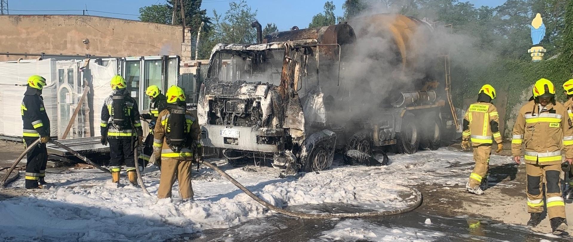 Po środku spalona betoniarka samochodowa. Po obu stronach pracujący strażacy stoją w pianie gaśniczej