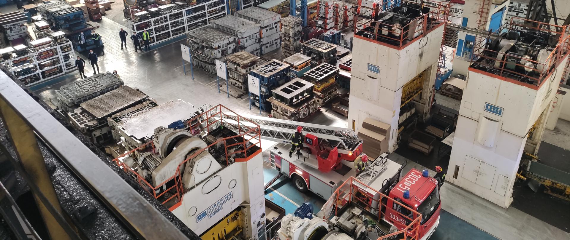 Zdjęcie przedstawia hale produkcyjną. Z lewej strony u góry widać suwnice. Na dole znajduje się samochód strażacki - drabina - przygotowuje się do rozstawienia.