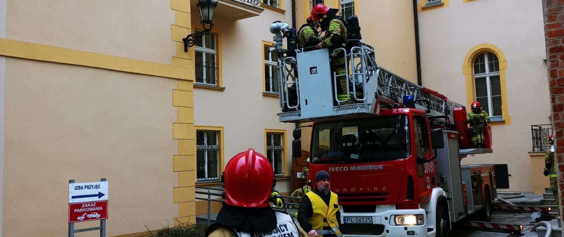 Drabina mechaniczna stoi przy budynku. W koszu wysięgnika strażacy.