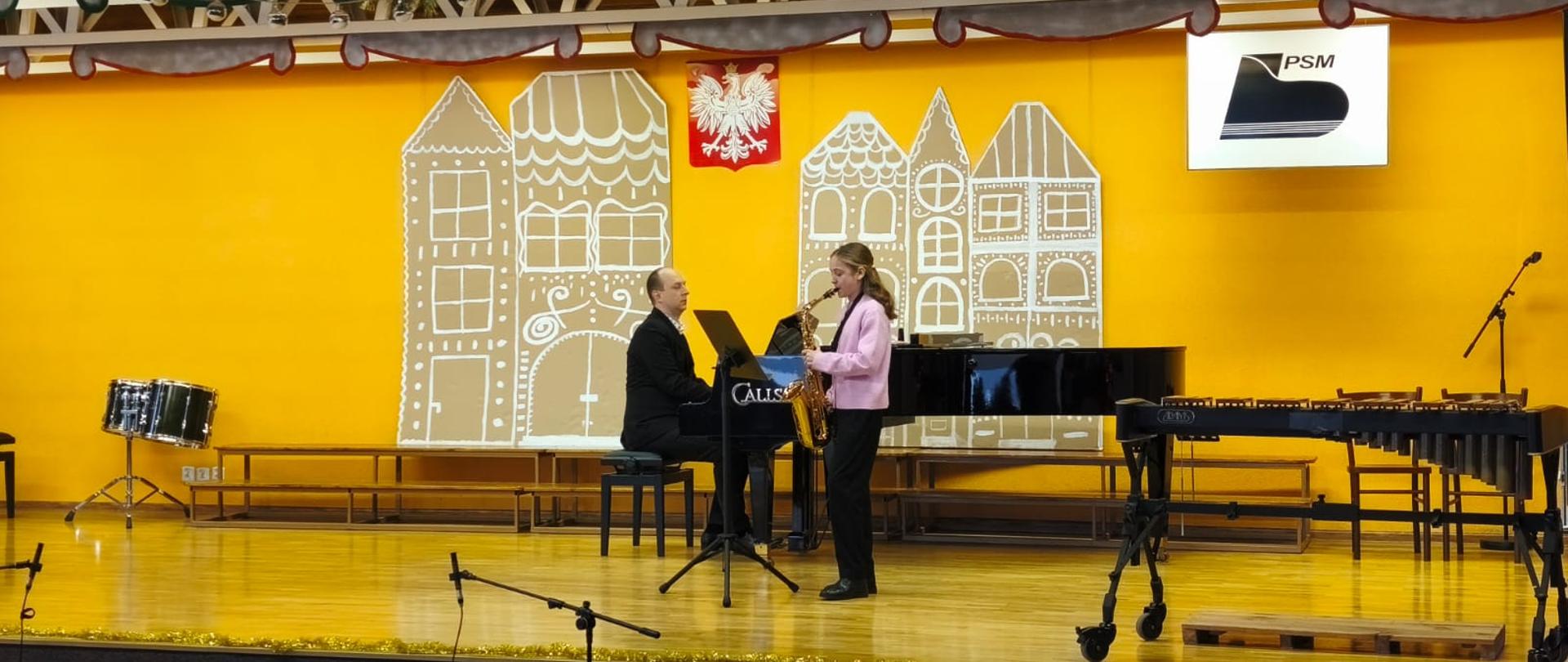 Uczennica gra na instrumencie. W tle czarny fortepian. Akompaniator. Żółta ściana. Ozdoby zimowe.