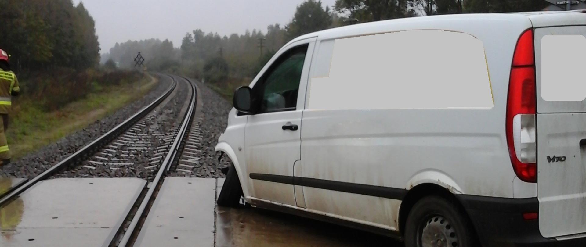 Uszkodzony samochód dostawczy na przy przejeździe kolejowym po zderzeniu z szynobusem. Uszkodzona przednia oś auta. W oddali ledwie widoczny tył szynobusa.