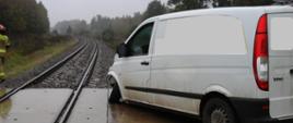 Uszkodzony samochód dostawczy na przy przejeździe kolejowym po zderzeniu z szynobusem. Uszkodzona przednia oś auta. W oddali ledwie widoczny tył szynobusa.