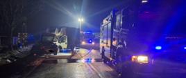Zdjęcie przedstawia dwa samochody po kolizji drogowej, samochód osobowy koloru czarnego oraz samochód dostawczy koloru białego z plandeką. Na drugim planie pojazdy pożarnicze podczas działań ratowniczych.