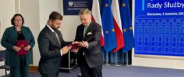 Prezydent Bełchatowa Patryk Marjan odbiera akt powołania z rąk min. Jakuba Stefaniaka, sekretarza stanu, Zastępcy Szefa Kancelarii Prezesa Rady Ministrów; w tle flagi Polski i Unii Europejskiej