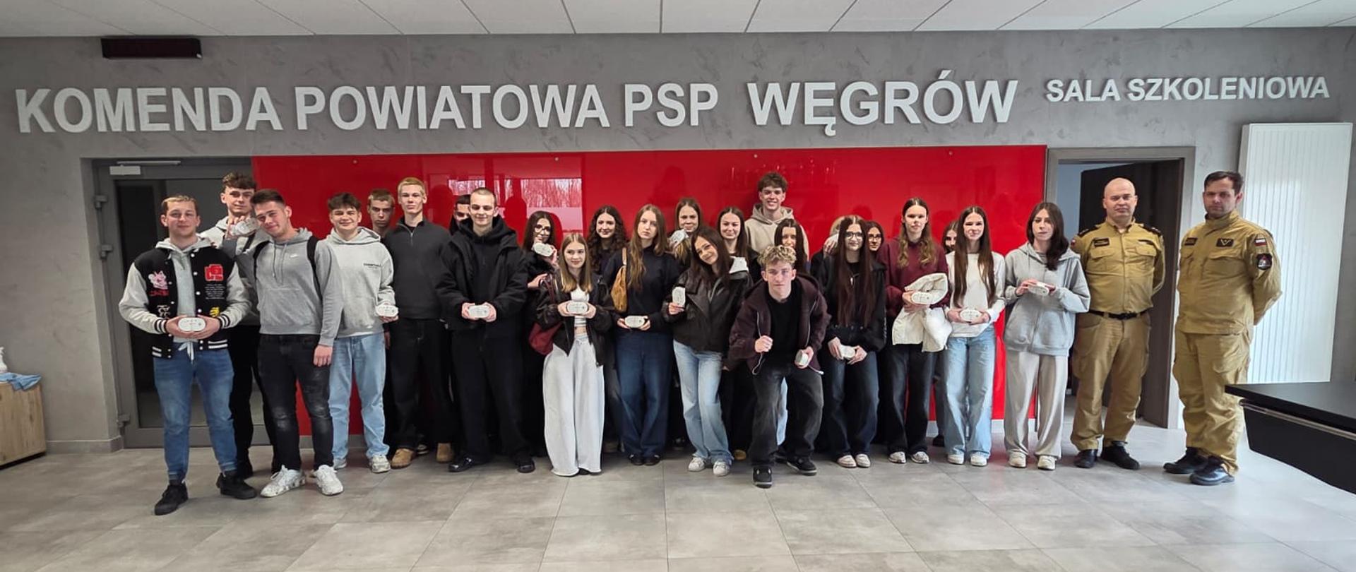 Na zdjęciu przedstawiono grupę młodzieży oraz strażaków podczas wspólnego spotkania w sali szkoleniowej Komendy Powiatowej Państwowej Straży Pożarnej w Węgrowie. Uczestnicy ustawieni są w jednym rzędzie na tle charakterystycznej ściany z napisem jednostki.
Młodzież trzyma w rękach czujniki dymu, co wskazuje na zakończenie zajęć edukacyjnych lub akcji profilaktycznej związanej z bezpieczeństwem pożarowym. Po prawej stronie widoczni są strażacy w umundurowaniu, którzy prawdopodobnie prowadzili szkolenie.
Całość przedstawia pamiątkowe zdjęcie podsumowujące spotkanie o charakterze edukacyjnym, mające na celu zwiększenie świadomości w zakresie ochrony przeciwpożarowej oraz promowanie zasad bezpieczeństwa wśród młodzieży.