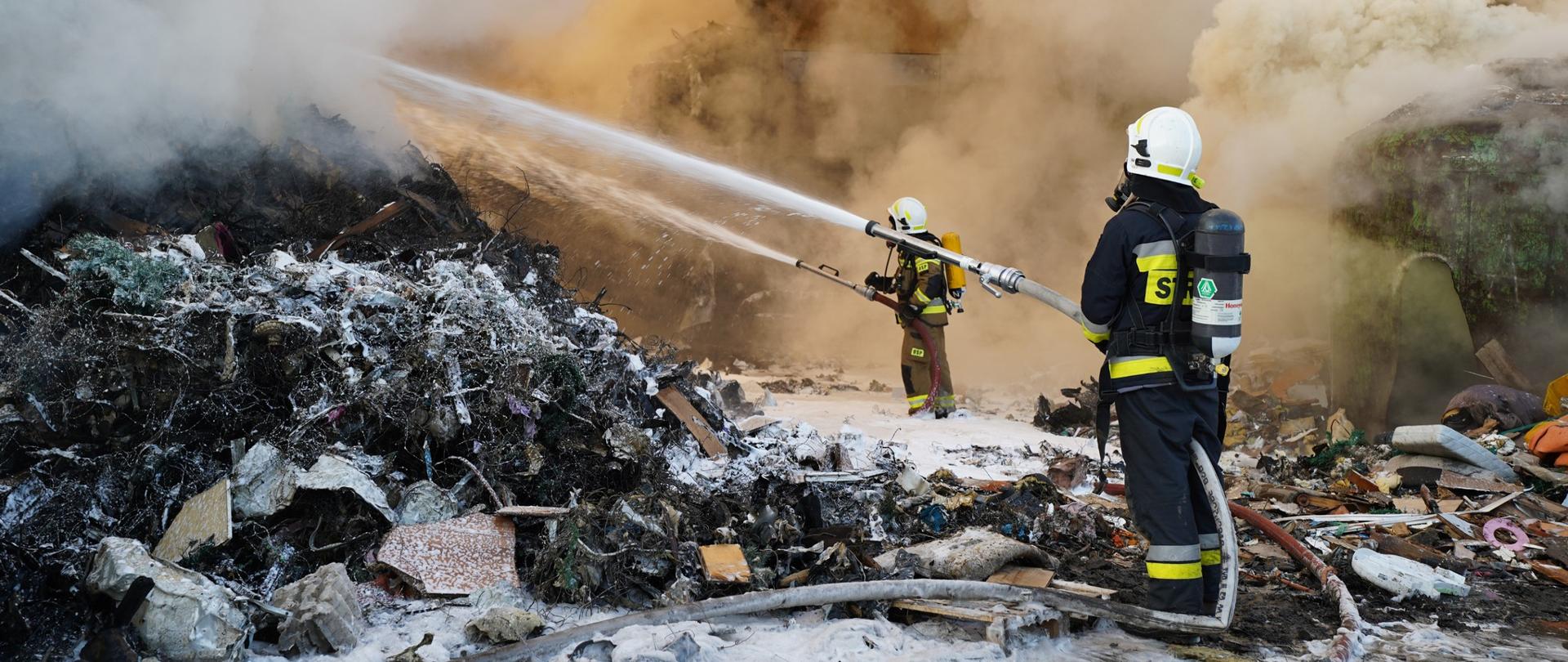 Strażak gasi pożar w zakładzie przetwarzania odpadów. Jest na samochodzie pożarniczym.