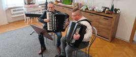 Na zdjęciu widać dwóch mężczyzn grających na czarnych akordeonach. Obaj siedzą na jasnych krzesłach. Przed nimi stoi pulpit na nuty. Mężczyzna siedzący z prawej strony jest ubrany w niebieskie spodnie oraz białą koszulę. Mężczyzna siedzący z lewej strony jest ubrany w niebieskie spodnie, białą koszulę oraz ciemną marynarkę. W tle widać brązową komodę z dekoracją świąteczną, czarny instrument klawiszowy na statywie.